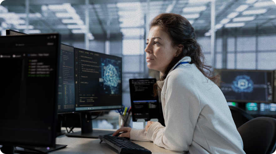 Alt text: A focused IT professional, a woman with dark hair, works at a multi-monitor computer station in a data center or tech environment. Her screens display lines of code and technical diagrams, symbolizing enhanced security and a proactive security posture.