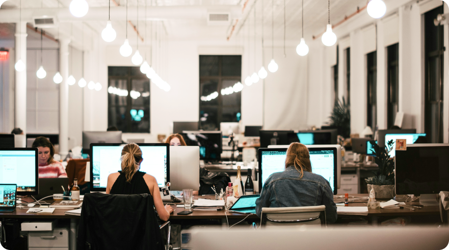 Alt text: An office workspace filled with employees sitting at desks with multiple computer monitors. The photo is taken from a slight distance and has a warm, vintage-like filter. The hanging lights are bokeh and there's a laptop with the ControlUp logo on one of the desks.
