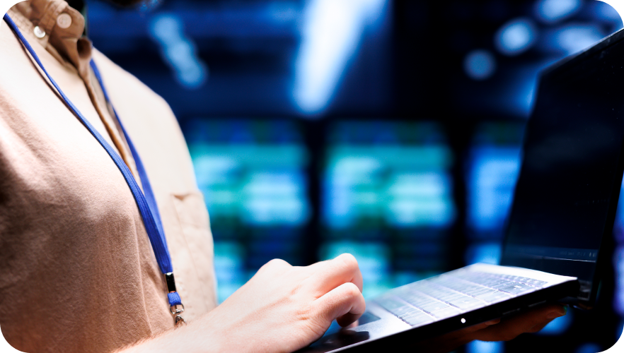 A close-up, side-view shot of a technician in a beige shirt holding a laptop in a server room. His hand rests on the keyboard, and the background is filled with the blurred blue lights of server racks.