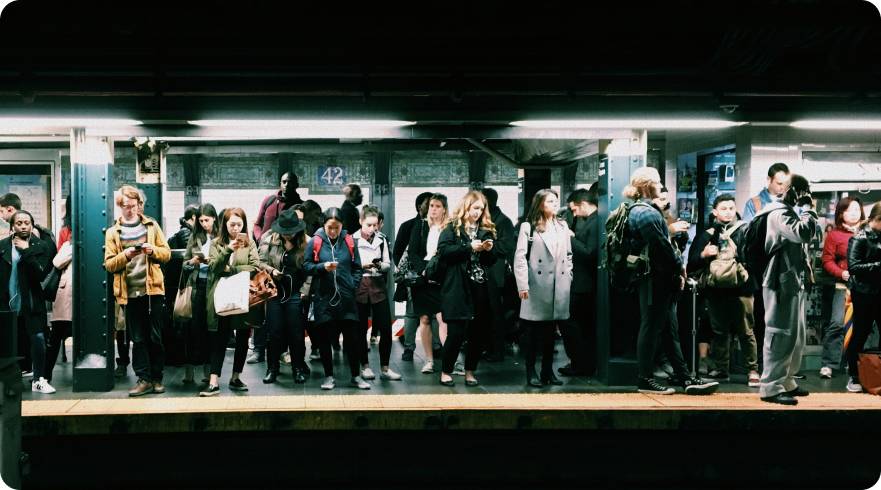 A wide-angle photo of a diverse crowd of people standing on a subway platform. Most individuals are looking down at their smartphones or other mobile devices. The scene is dim, lit by fluorescent lights. This image represents the concept of **Mobile Device Management** (MDM) and the widespread use of mobile devices in a public setting.